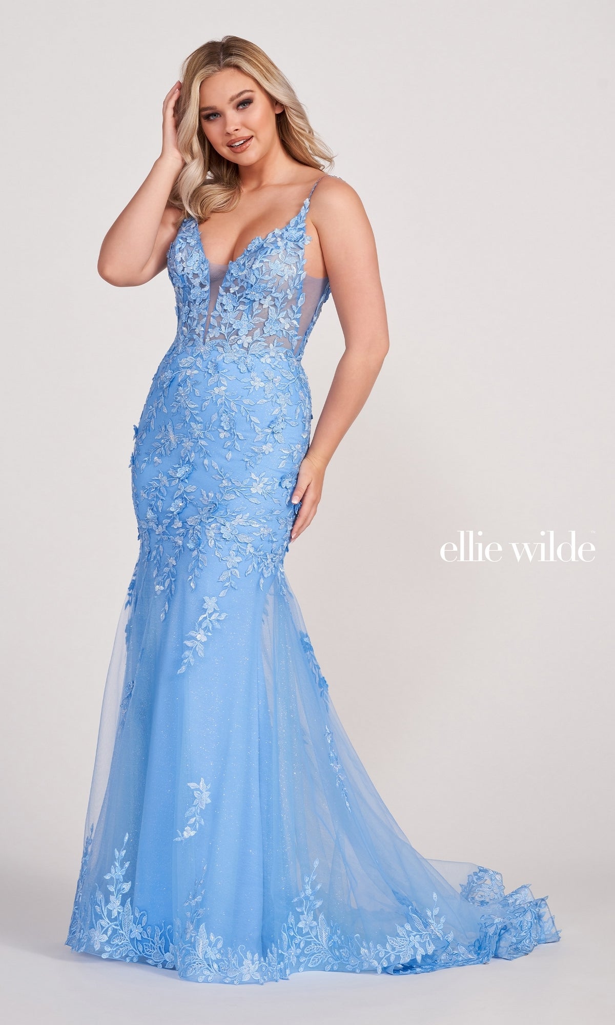 A woman poses against a plain background in a Lace Embroidered Mermaid Gown With Sheer Bodice, showcasing floral appliqué, sheer panels, and a light blue fitted silhouette.